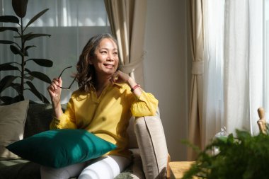 Orta yaşlı bir kadın rahat koltukta dinleniyor, başka yere bakıyor, hayal kuruyor stresten uzak, huzurlu bir ruh halinin keyfini çıkarıyor oturma odasında yalnızken..