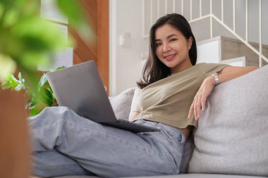 Evde rahat bir kanepede dinlenirken dizüstü bilgisayar kullanan çekici genç bir kadın..