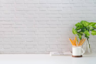 Boş resim çerçevesi, kahve fincanı, kitap ve kırtasiyesi beyaz masada, tuğla duvara dayalı rahat bir iş yeri. Metnin için boşluk kopyala.