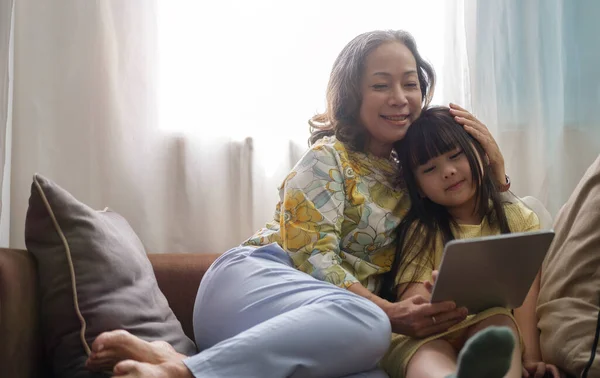 Happy grandma and cute little girl sitting on sofa and watching cartoons on digital tablet together.