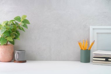 Stylish workplace with picture frame, coffee cup, potted plant and supplies on white table. Copy space.	