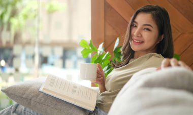 Oturma odasındaki kanepede oturmuş, evde boş vaktinin keyfini çıkarırken çay içip kitap okuyan güzel genç bir kadın..