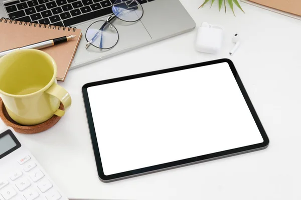 Digital tablet with blank screen, laptop computer, calculator and coffee cup on white table.