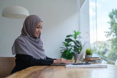 Gülümseyen, tesettürlü genç iş kadını modern ofiste dizüstü bilgisayarla çalışıyor..