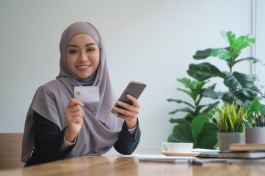 Mutlu İslamcı iş kadını elinde kredi kartı tutarken akıllı telefon üzerinden online bankacılık yapıyor. Çevrimiçi alışveriş, e-ticaret kavramı.
