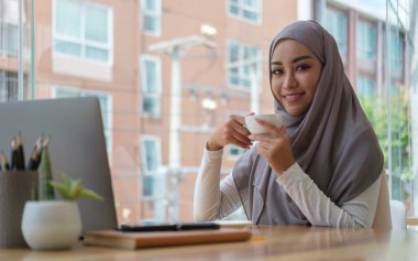 Güzel Müslüman iş kadını yönetici tesettür giyiyor sıcak kahve içiyor ve modern ofiste çalışıyor..