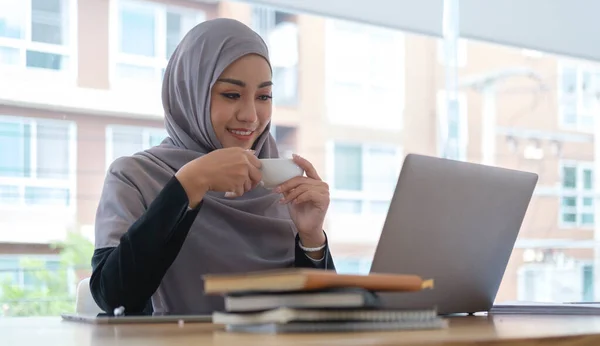 Happy muslim businesswoman in headscarf drinking hot coffee and reading online news on laptop.