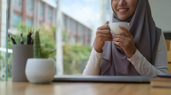 Smiling muslim businesswoman boss manager enjoy her morning coffee, sitting near large window with city buildings in background.