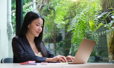 Genç Asyalı kadın ofis çalışanı parlak ofiste oturuyor ve bilgisayar bilgisayarı üzerinde çalışıyor..