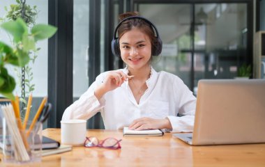 Kulaklık takan ve bilgisayar bilgisayarı üzerinde çalışan mutlu, genç bir kadın..