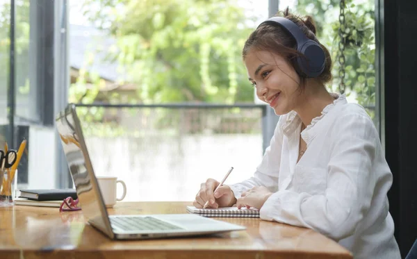 Smiling young woman wearing headphone, studying online course via laptop and lecture on notepad.