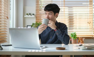 Düşünceli Asyalı genç bir adam kahve fincanı tutuyor ve modern ofiste otururken pencereden dışarı bakıyor..