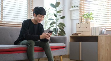 Genç Asyalı adam kanepede dinleniyor ve akıllı telefon kullanıyor..