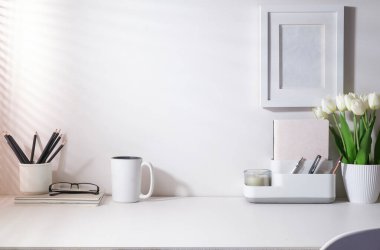 Kahve fincanı, kırtasiye ve çiçek saksısı beyaz masanın üzerinde güneş ışığı pencerenin perdelerinden geçiyor..