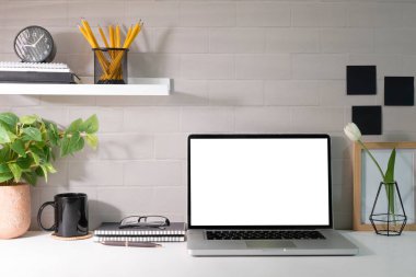 Laptop white empty screen, picture frame, potted plant and supplies on white table. Stylish workplace.