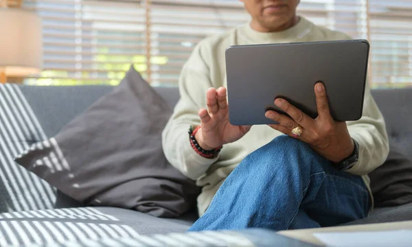 Retired Asian man resting on couch and using digital tablet for browsing internet or reading news online in the morning.