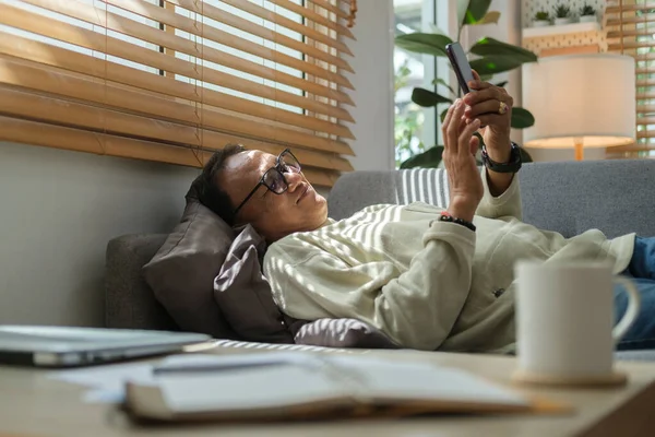 Relaxed middle aged man lying on couch and using smart phone. Older people and gadgets.