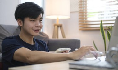 Gülümseyen yakışıklı Asyalı serbest yazar dizüstü bilgisayarla çalışıyor..