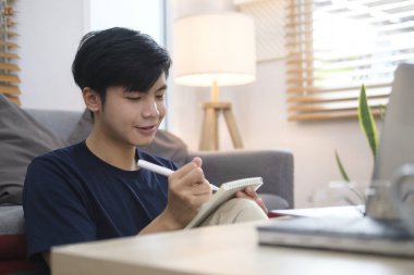 Young asian man sitting in bright living room and working online with laptop.