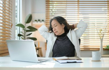 Satisfied mature businesswoman holding hands behind head taking break form work and relaxing at her workplace.