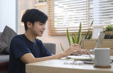 Gündelik giysiler içinde gülümseyen Asyalı adam dizüstü bilgisayar kullanıyor, internette geziniyor ya da evde bilgisayarla çalışıyor..