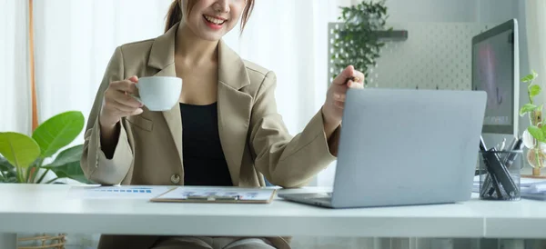 Millennial businesswoman drinking hot coffee and reading email or online news on her laptop computer.