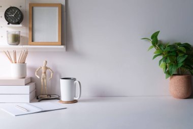 Comfortable workplace with houseplant, picture frame, coffee cup and stationery on white table.