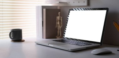 Computer laptop with empty screen, books, coffee cup and pencil holder on white table in bright living room.