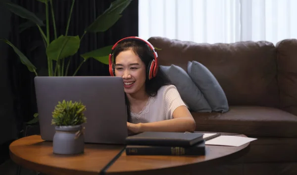Cheerful young woman in wireless headphone surfing internet on laptop, while sitting in cozy living room. E-learning education concept.