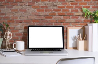 Öndeki dizüstü bilgisayar, kırtasiye malzemesi, resim çerçevesi, ev bitkisi ve kahve fincanı beyaz masada. Şık bir iş yeri.