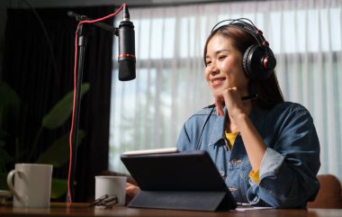 Smiling young woman in headphones using microphone and digital tablet broadcasting her live audio podcast from home studio.