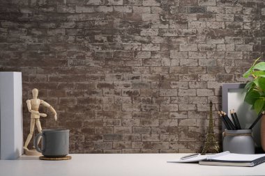 Creative workspace with blank poster frame, coffee cup, books and flower pot on white table with brick wall.	