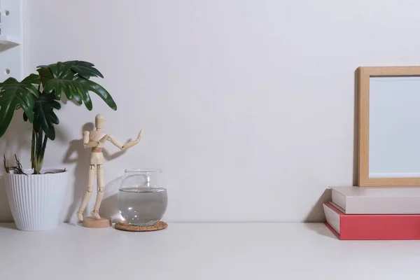 Glass of water, empty picture frame, books and houseplant on white table. Home office desk.