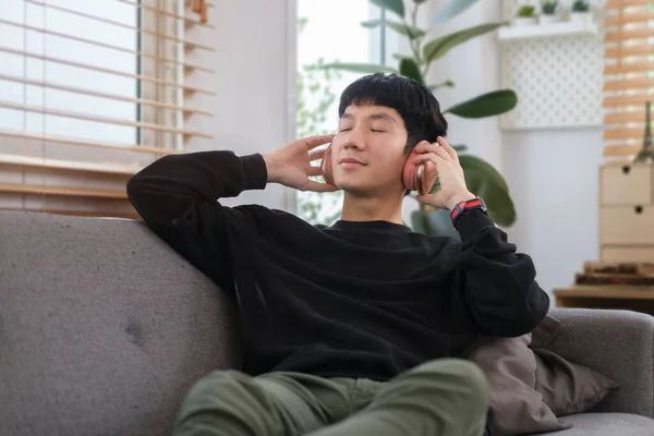 Relaxed man wearing warm sweater listening to music in wireless headphone and lying on couch at home.
