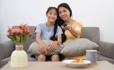 Asyalı bir anne elinde uzaktan kumandayla evde sevgili kızıyla televizyon izliyor, hafta sonu aile eğlencesi..