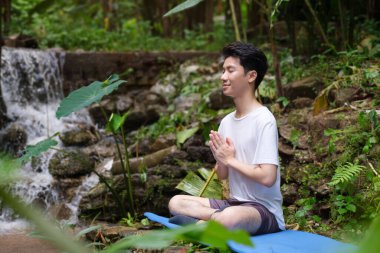 Tatmin olmuş, gözleri kapalı, meditasyon yapan ve şelalenin yanındaki ormanda kayalarda oturan bir adam. Yoga, pilates, sağlıklı yaşam tarzı konsepti.