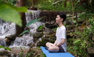 Genç adam yağmur ormanlarındaki şelalenin yakınındaki kayalıklarda meditasyon yapıyor ve oturuyor. Spor, yoga, pilates, sağlıklı yaşam tarzı.