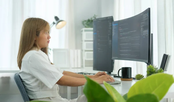 Focused female software developer working with program code on wide displays at modern workstation.