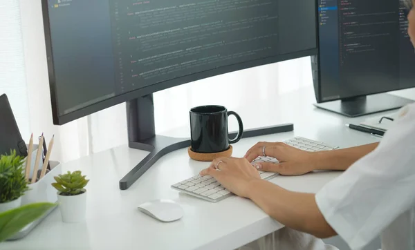 Cropped shot of young programmer writing program code with two monitors in bright office.