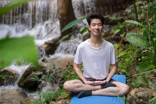 İnsan meditasyon yapıyor, yağmur ormanlarındaki şelalenin yakınındaki kayalıklarda oturuyor. Spor, yoga, pilates, fitness, sağlıklı yaşam tarzı konsepti.