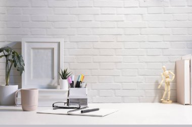 Ön manzara çerçevesi, kitaplar, kahve fincanı, tuğla duvara karşı beyaz masadaki kırtasiye..