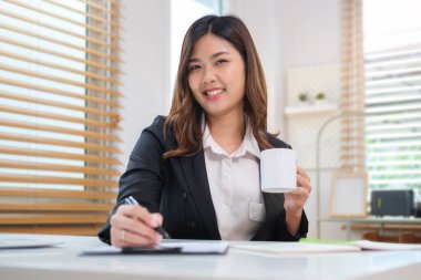 Gülümseyen genç iş kadını elinde kahve fincanı tutuyor ve belgedeki mali bilgileri kontrol ediyor..