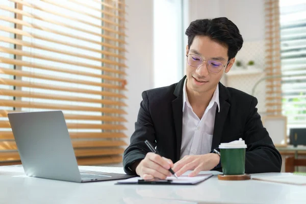 专注的亚洲商人在笔记本上写下重要信息 并在工作台上使用笔记本电脑 — 图库照片