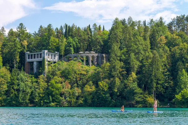Lake Bled, Slovenia - August 20, 2018: A couple relax on turquoise waters while paddle surfing in front of a lush green forest