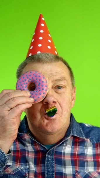 Senior volwassen blanke man met een verjaardagspet past multi-gekleurde zoete donuts op zijn ogen. — Stockvideo