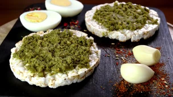 Rice cake toasts with spreading green pesto sauce and boiled eggs rotate slowly on cutting board. — ストック動画