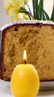 Evde yanan mum ve penceresinde taze nergis bulunan geleneksel Paskalya pastası dilimlenmiş. Geleneksel tatil yemekleri. Dikey. Yakın plan..