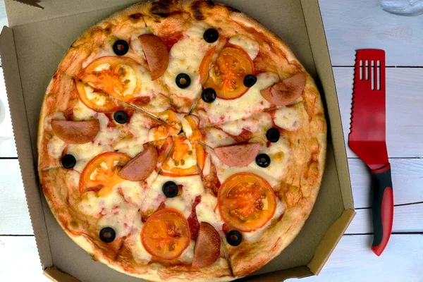 Fresh round pizza with tomatoes, sausage, mozzarella and olives in cardboard box. Pizza cutter and pizza spatula. Tasty unhealthy fast food. Close-up.