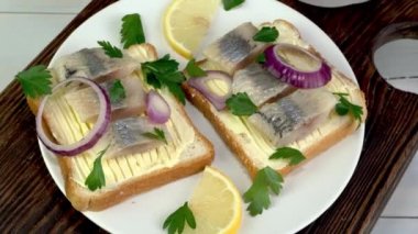 Tasty sandwich with salted herring fish fillet with onion rings, lemon and parsley rotate in plate. Traditional healthy snack. Close up.