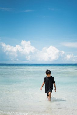 Asyalı çocuk tatillerde plajda oynuyor. Doğada deniz, kum ve mavi gökyüzü olan bir çocuk. Deniz kenarında tatilde olan mutlu genç çocuk suda yürüyor..